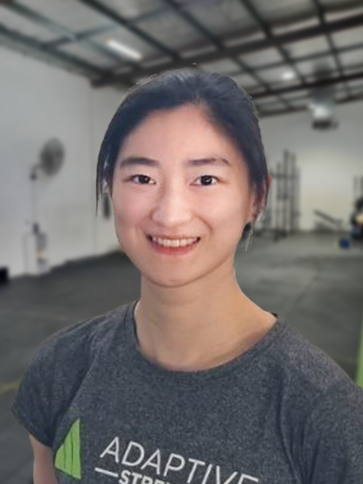 Physio_Profile_Prescilla31 - Adaptive Physiotherapy & Massage - Adaptive Physiotherapy & Massage Smiling woman with dark hair wearing a gray shirt that reads ADAPTIVE, in a spacious indoor workshop setting.