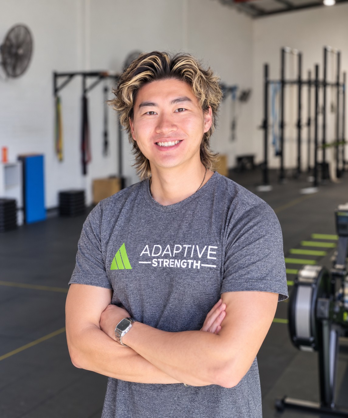 Smiling fitness trainer with arms crossed in a gym, wearing a gray Adaptive Strength shirt.