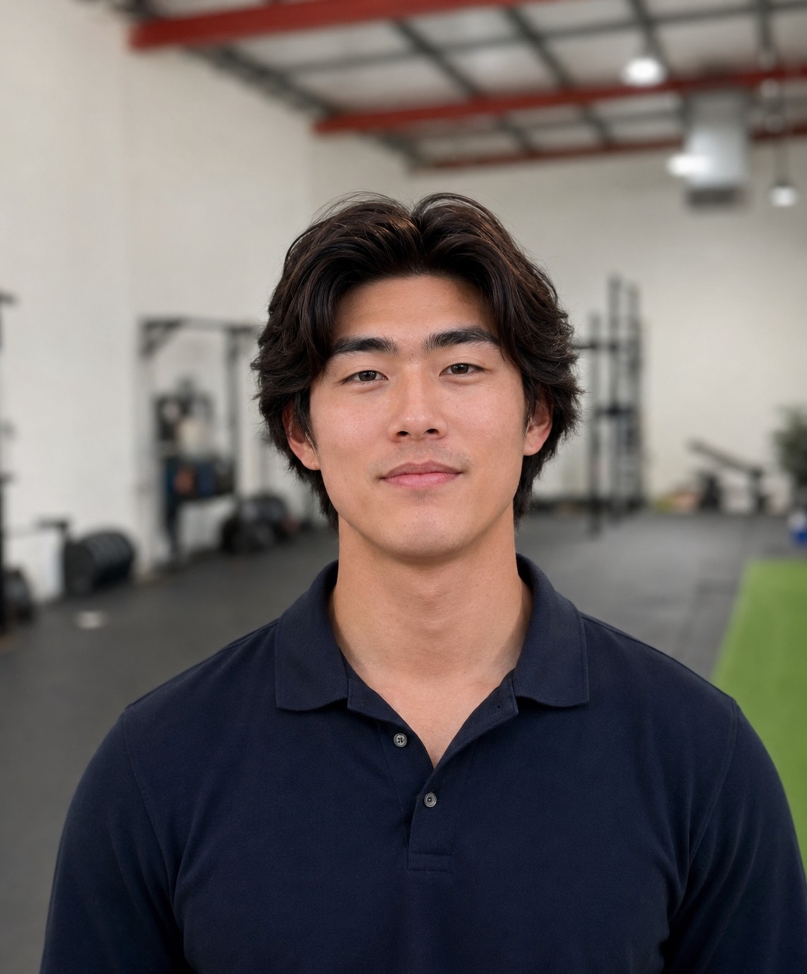 Portrait of a young man with dark hair wearing a navy polo inside a gym or fitness facility, looking at the camera with a neutral expression.